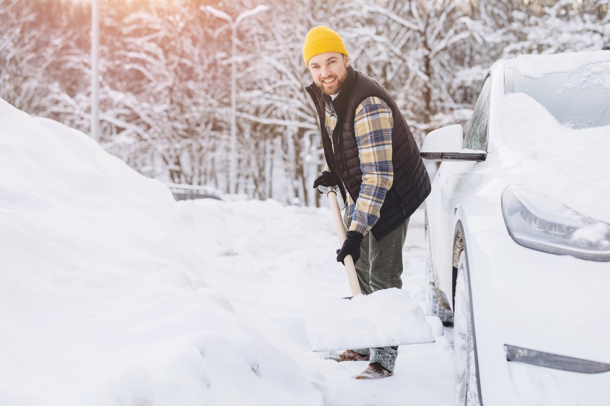 Man actively shoveling snow from a private driveway next to a white car, clearing a path after a heavy winter snowfall