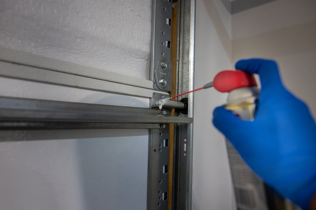 Regular garage door maintenance is important to reduce friction, wear, noise and rust. Homeowner applies lubricant to wheel bearings on door